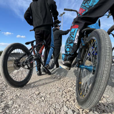 Kaksi polkupyörää muistuttavaa BMX-pyörää kuskeineen kuvattuna takaapäin. Taustalla sininen taivas.