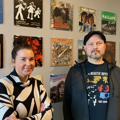 Näyttelyamanuenssi Elina Suutari ja näyttelyn kuratoinut levykauppayrittäjä Ari-Pekka Savolainen seisovat seinän edessä, jonne on ripustettu levynkansia. 