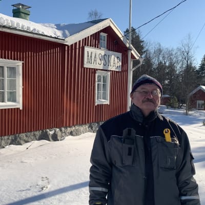 En man i vinterkläder står framför en gammal träbyggnad i snö. 