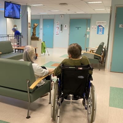 Personer i väntrummet vid en hälsocentral, en person i rullstol längst fram. 