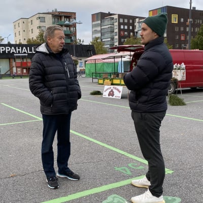 Kaksi aikuista miestä seisoo torilla, taustalla näkyy kauppahallin rakennus.