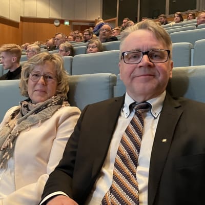En man i kostym  och en kvinna i rosa dräkt sitter längst fram i ett halfullt auditorium.