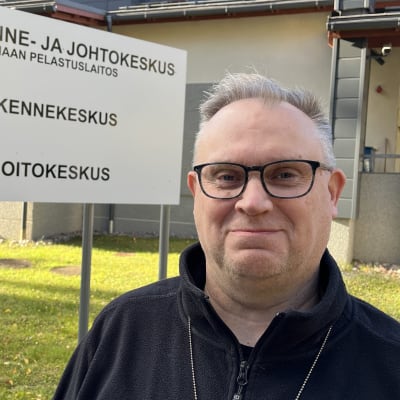 Mustaan takkiin pukeutunut, harmaita hiuksia päälaella omaava mies katsoo kuvaajaa ja hymyilee hiukan. Kuvattu ulkona, aurinko paistaa, taustalla rakennus ja kyltti, jossa kukaa mm, ensihoitokeskus. 