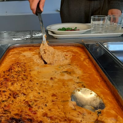 En sked sticks ner i en stor form med orange brolierpasta i en skolmatsal.