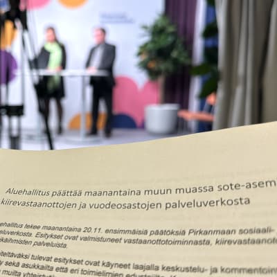 Etualalla on paperi, jossa kerrotaan aluehallituksen päättävän maanantaina sote-asemien, kiirevastaanottojen ja vuodeosastojen palveluverkosta. Taustalla on epätarkasti ihmisiä ja kameroita.