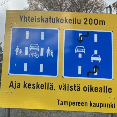 Iso keltainen liikennemerkki, jossa teksti "Yhteiskatukokeilu 200m. Aja keskellä, väistä oikealle. Tampereen kaupunki". Kyltissä myös sinisellä pohjalla havainnoillistavat kuvat
