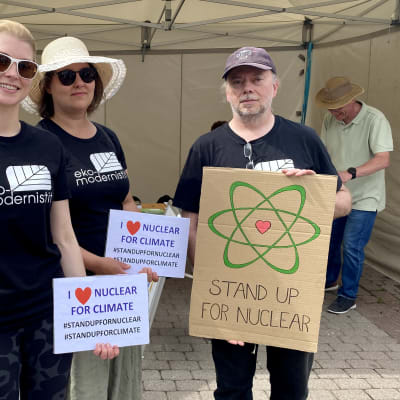 Personer vid torgtält håller upp skyltar för kärnkraft, bland annat med texten Stå upp för kärnkraft.