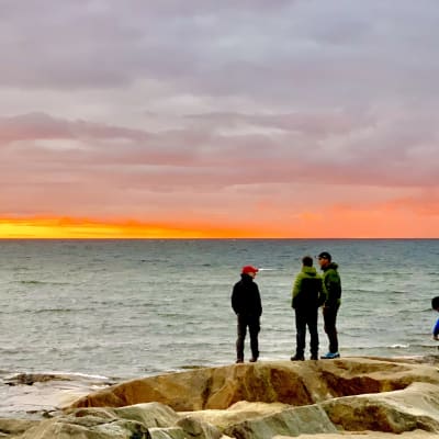Tre personer står på klipporna vid havet i solnedgången.