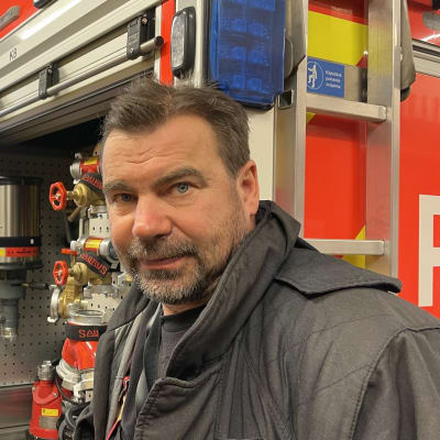 Palomies Juhani Prokki katsoo kameraan, taustalla paloauto.
