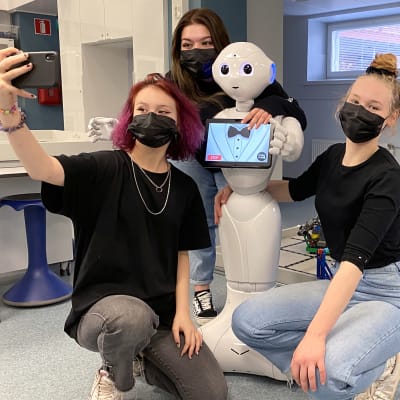 Jenna Hietakangas, Pinja Jokinen ja Sara Korkia-aho ottavat selfietä yhdessä ihmishahmoa muistuttavan humanoidirobotin kanssa.