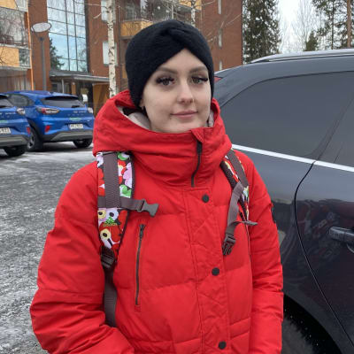 Punaiseen takkiin pukeutunut Sari Toivonen seisoo tumman auton vieressä kädet takin taskuissa ja katsoo kameraan.