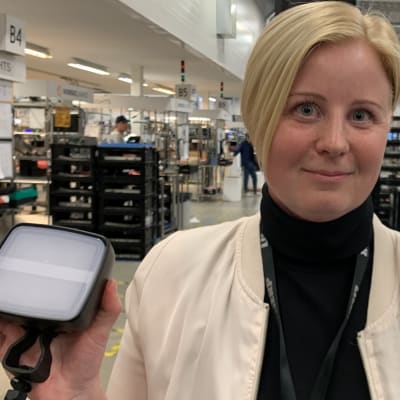 En kvinna med vit blazer står i en fabrikshall och håller upp en lampa i ena handen. 