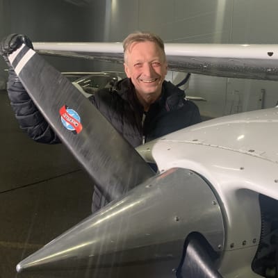 En glad man står och håller i propellern på ett småplan i en flyghangar