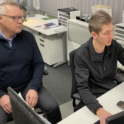 Två män sitter vid ett bord och tittar på en datorskärm.
