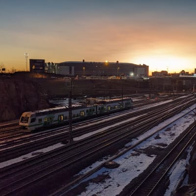Ett tåg kör i ett vintrigt landskap i Ilmala. I bakgrunden en stigande sol och Helsingfors Arenan.