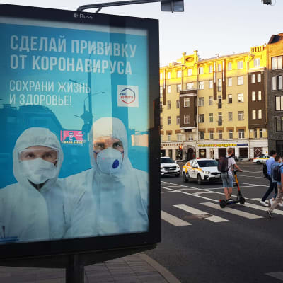Moskvaborna uppmanas ta coronavaccin. En skylt i stadsbilden kommer med uppmaningen. I bakgrunden går folk över ett övergångsställe.