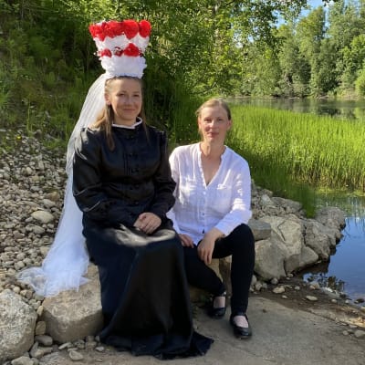 Kaksi naista istuu jokirannassa. Toisella on kruunuhäiden morsiamen asu päällään: musta pitkä puku, valkoinen huntu ja korkea koristeltu kruunu.