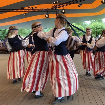 Kansallipukuisia lapsia tanssimassa Kaustisen Kansanmusiikkijuhlien pääareenalla. Mukana tyttöjä ja poikia vauhdikkaassa tanssissa.
