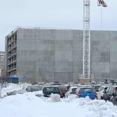 Lahden betoninen pysäköintilaitos rakenteilla Ranta-Kartanossa, korkea nosturi rakennuksen etu-alalla, autoja edessa pysäköitynä, lunta maassa.
