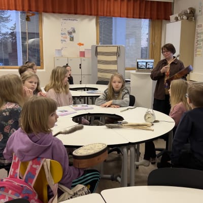 Opettaja Sari H. Niemelä seisoo rumpu kädessään ja oppilaat istuvat kahden pyöreän pöydän ympärillä. 