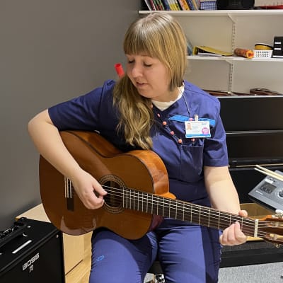 Siniseen sairaalan työasuun pukeutunut nainen istuu kitara kädessään