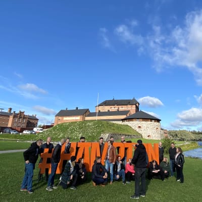 Joukko ihmisiä ryhmittäytyy oranssisten Hämpton-kirjaimien luo puistossa. Takana vanha linna, sinitaivas.ja järvi.