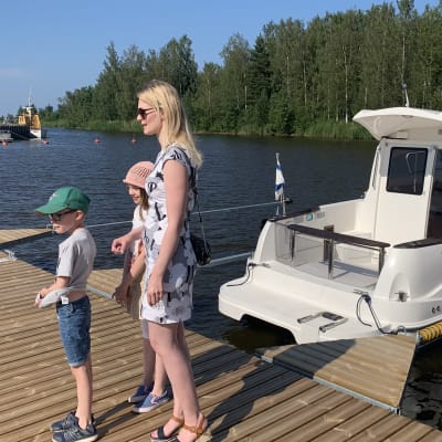 En kvinna går på en brygga med småbåtar, i sällskap av en flicka och en pojke. 