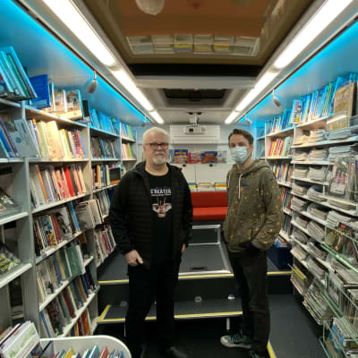 Kirjastoauton käytävällä seisoo kaksi miestä. Käytävän laidoilla on paljon kirjoja hyllyissä, takaosassa on punainen sohva.