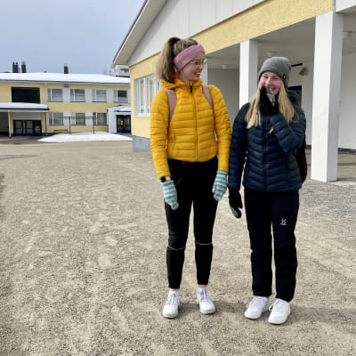 Kaksi nuorta tyttöä seisoo koulurakennuksen edessä ja toista naurattaa niin, ettähän on nostanut käden suunsa eteen.