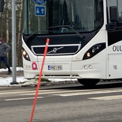 Oulun joukkoliikenteen valkoinen bussi ajaa tiellä. Taustalla jalankulkija kävelee jalkakäytävällä.