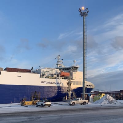 Ett stort containerfartyg står vid hamnkajen med bilar och arbetsmaskiner i förgrunden. 