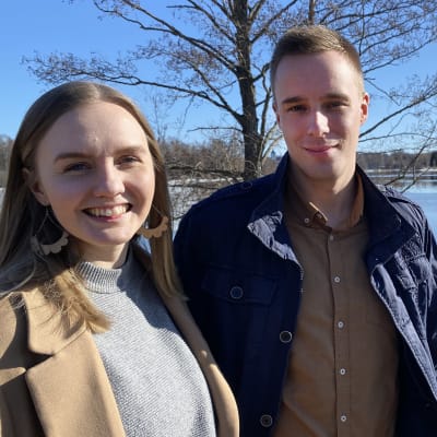 Laura Saartoala ja Jari Mäkäläinen seisovat järven rannalla kevätauringossa takanaan lehdetön puu ja vastarannalta näkyvä Hämeen vanha linna.