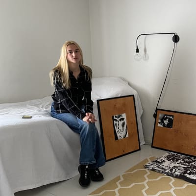 Kvinna som sitter med benen i kors på en säng, omgiven av konst.