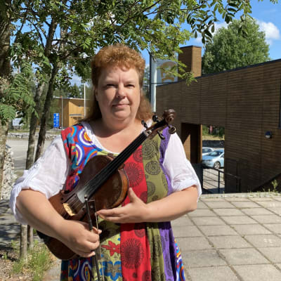Anneli Aho pitelee viulua käsissään ja on pukeutuneena värikkääseen esiintymisasuun. Taustalla on Kaustisen Kansantaiteenkeskuksen rakennus