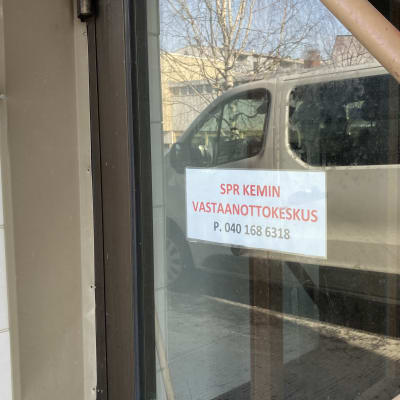 Ulko-ovi, jonka ikkunassa on lappu ja teksti SPR Kemin vastaanottokeskus. Ikkunasta heijastuu iso valkoinen pakettiauto.