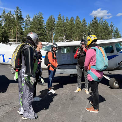 Viisi Hämeen laskuvarjourheilijoiden hyppääjää astumassa lentokoneen sisuksiin Lahti-Vesivehmaan lentokentällä. Sininen taivas, kesäinen päivä.