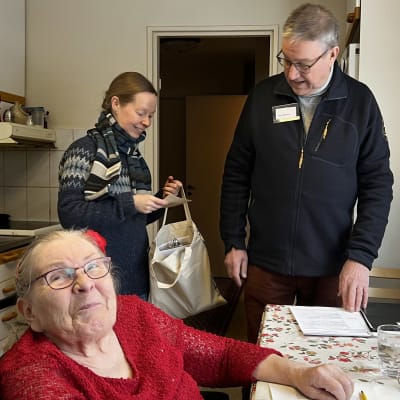 Lahtelainen Ritva Aittasalo äänestää kotona, istuu pyörätuolissa, katsoo kameraan ja hymyilee. Vieressä vaalitoimitsijat Reetta Rautiainen ja Niko Niemi.
