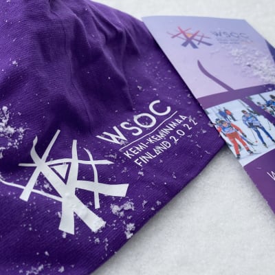 Hangen päällä on violetti pipo, jossa on WSOC logo ja teksti Kemi-Keminmaa sekä esite, jossa on teksti World Ski Orienteering Championships 2022.