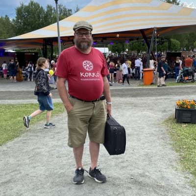 Jaakko Ruuttunen punaisessa t-paidassa seisoo soitinkoteloineen pelimannitalon rinteessä Festivaali-alueella. Taustalla konsertti areenalla.