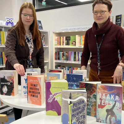 Katja Malmi ja Ulla Karila sijoittelevat kirjoja esille kirjaston pöydälle, taustalla kirjahyllyjä.