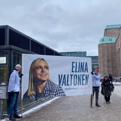 Två kampanjarbetare håller upp banderoll för Elina Valtonen framför järnvägsstationen i Helsingfors.