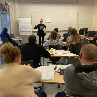 Luokka, jossa etualalla aikuiset suomenkielen opiskelijat istuvat selin, ja opettaja puhuu luokan edessä.