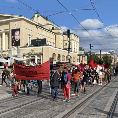 Ihmisiä kulkemassa jonomuodostelmassa kadulla. Mielenosoittajilla on käsissään kylttejä ja punaisia lippuja. Etualalla olevilla on kädessään iso punainen banneri.