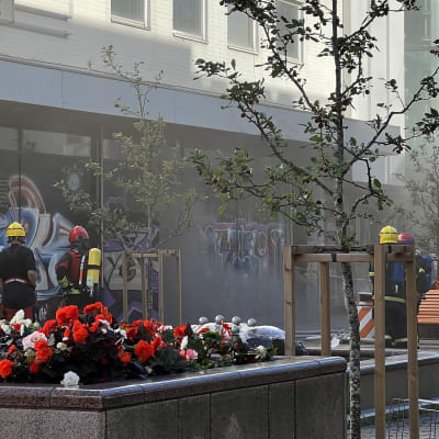 Kelta- ja punakypäräisiä palomiehiä savuavan rakennuksen vieressä kävelykadulla. Kuvassa kukkaistutuksia etualalla sekä muutama nuori puu ja penkki oikealla.