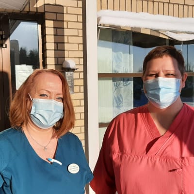 Två hälsovårdare med munskydd vid ingången till en hälsocentral, Carola Silander-Lehtinen i blå sköterskedräkt och Camilla Rautanen i röd sköterskedräkt.