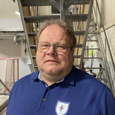 Silmälasipäinen mies seisoo jääkiekkomaalien vieressä. Hänellä on päällään sininen paita, jossa on jääkiekkojoukkueen logo.
