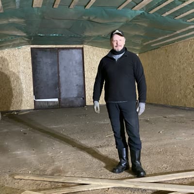 Mies seisoo tyhjässä tallirakennuksessa ja katsoo kameraan.