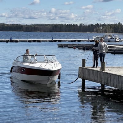 Mies moottoriveneessä Vääksyn Majakkapaviljongin satama-alueella kiinnittymässä laituriin. Laiturilla mies ja nainen pitävät kiinni veneen köysistä.