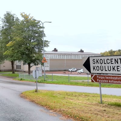 En vit vägskylt med texten Skolcenter Koulukeskus, och en brun skylt med texten Pargas hembygdsmuseum, i en vägkorsning med en skolbyggnad i bakgrunden.