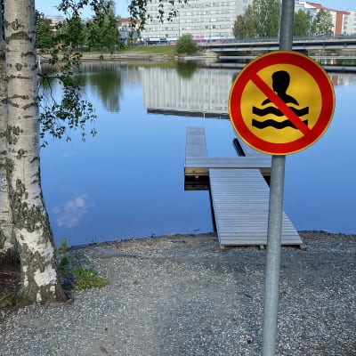 Kuvassa Limppusaaren uintikieltomerkki, jonka taustalla laituri vedessä. Horisontissa näkyy Kajaanin keskustaa.